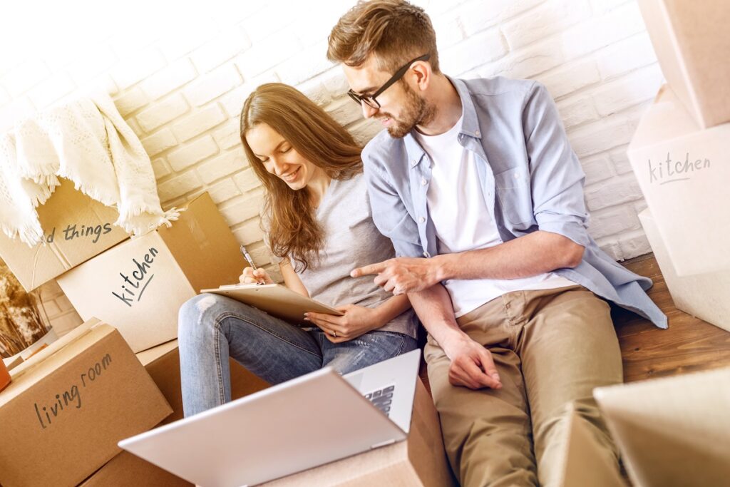 a couple sitting on the ground, looking at a tablet, surrounded by boxes