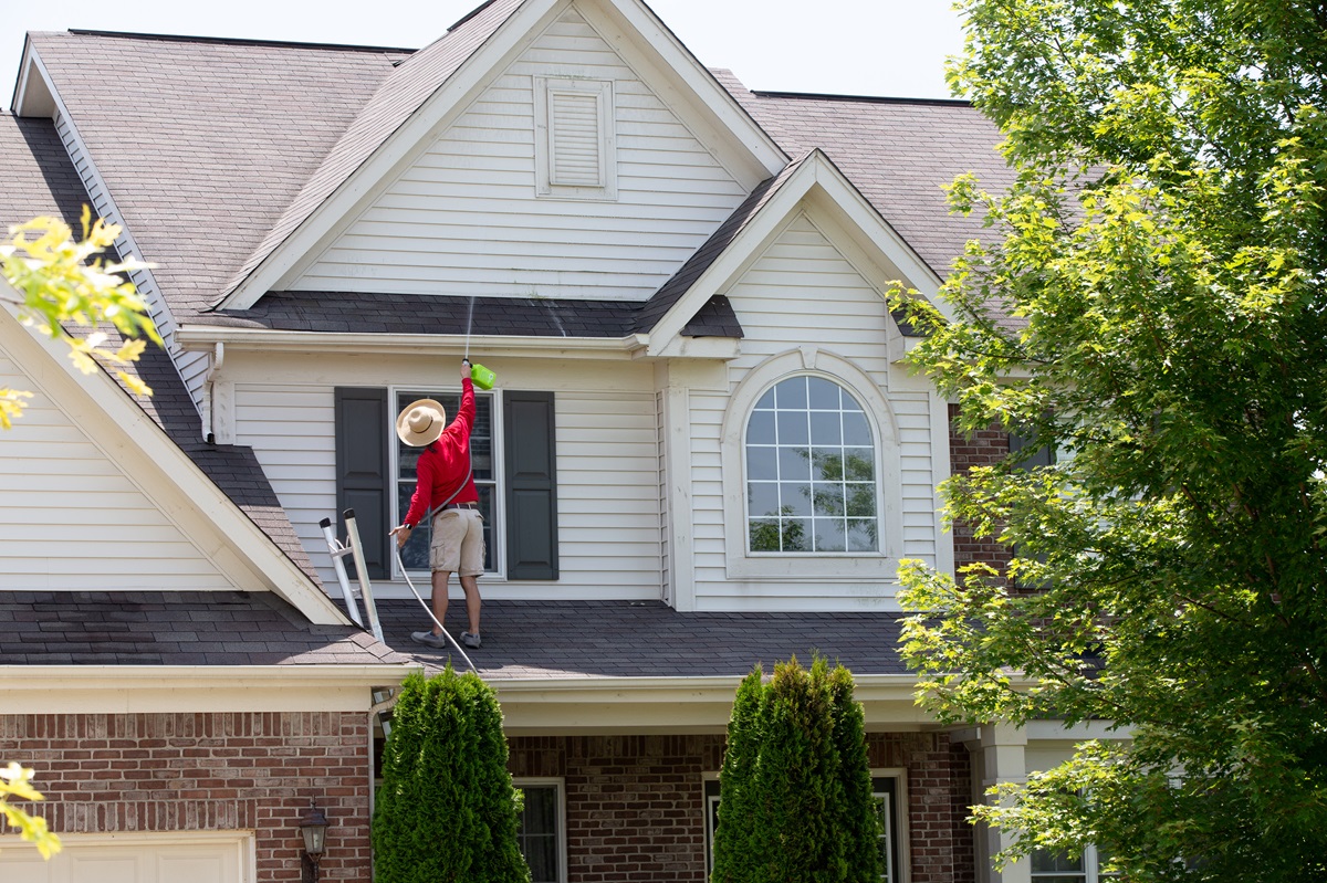 Homeowner washing the exterior of his house in spring balancing on the lower roof to spray the second floor facade with a handheld pressure sprayer