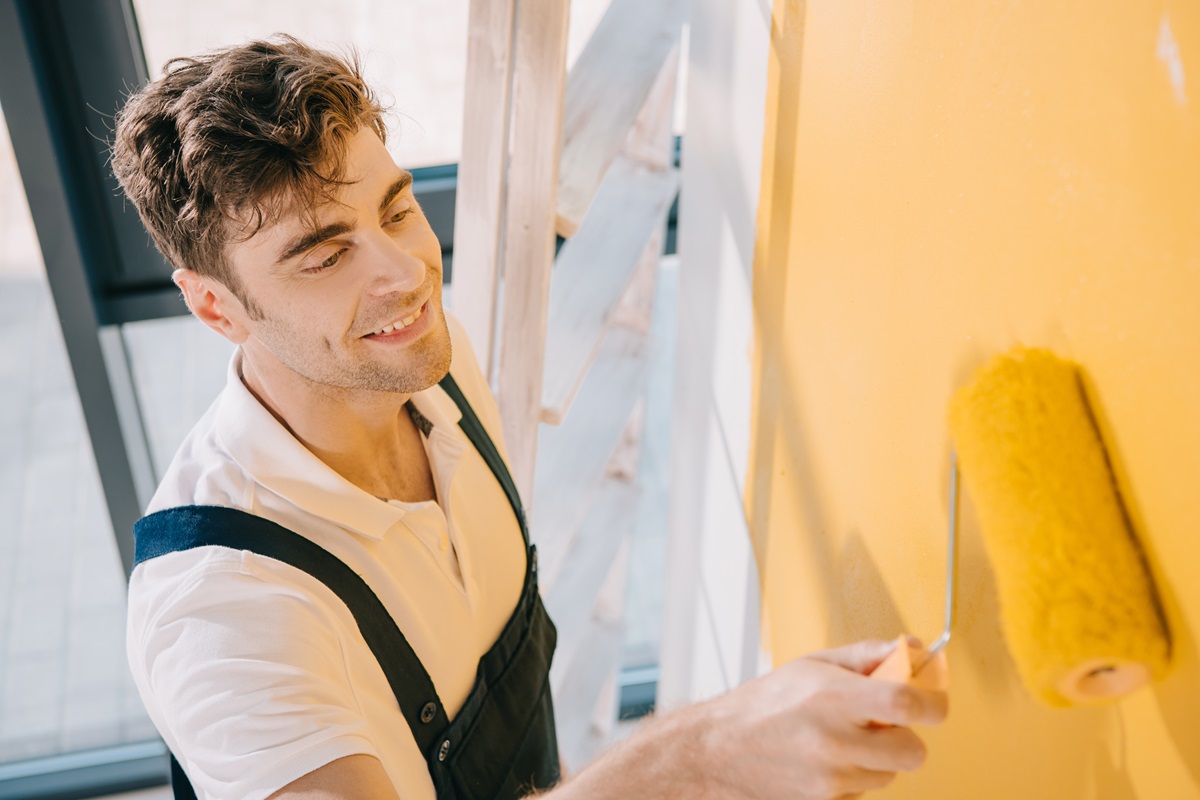 young painter smiling while painting wall in yellow color with paint roller