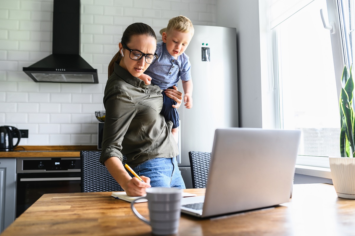 a busy woman with a child on her hip in her kitchen in front of a computer while writing something on a notepad